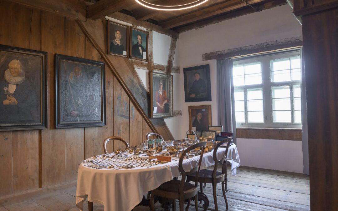 Ein Raum mit Holzboden, Holzdecke und Fachwerk-Wänden; an den Wänden alte Porträts, in der Mitte der Raums ein Tisch mit einem weissen Tischtuch, darauf diverses Silberbesteck